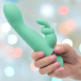 A hand with pink-painted nails holds the CalExotics Love Bunny Rockin’ “G” Bunny, a green silicone rabbit-style vibrator, against a softly blurred pastel background.